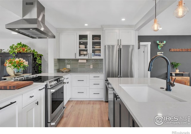 kitchen with appliances with stainless steel finishes, wall chimney range hood, a sink, light countertops, and glass insert cabinets