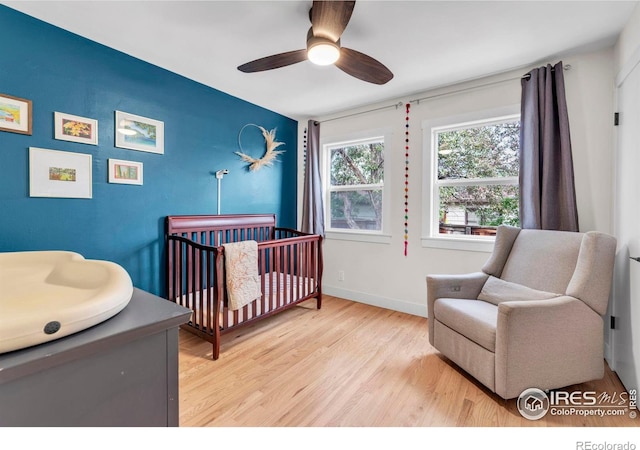 bedroom with a nursery area, light wood-style floors, an accent wall, baseboards, and ceiling fan