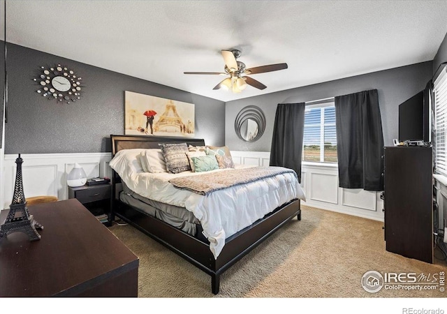bedroom featuring a decorative wall, wainscoting, carpet, and ceiling fan