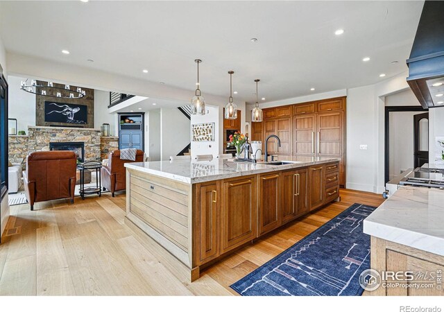 kitchen with light wood finished floors, a sink, light stone countertops, a stone fireplace, and a large island with sink