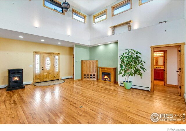 unfurnished living room featuring a glass covered fireplace, light wood-style floors, a wood stove, baseboard heating, and a high ceiling