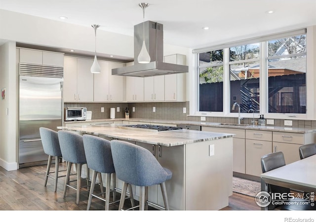 kitchen with built in refrigerator, island range hood, gas stovetop, a sink, and wood finished floors