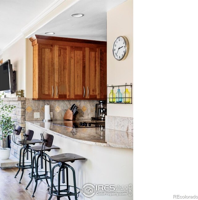 kitchen with a breakfast bar, decorative backsplash, brown cabinetry, light wood-style floors, and light stone countertops