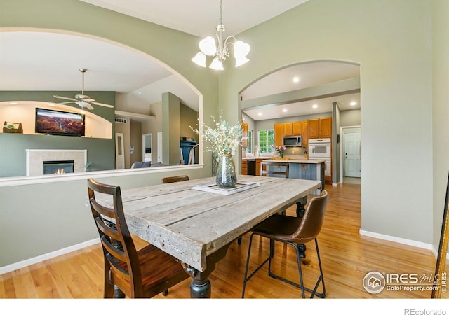 dining room featuring a tiled fireplace, a ceiling fan, light wood-style flooring, a chandelier, and lofted ceiling