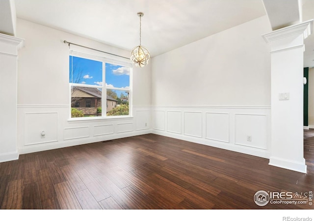 unfurnished dining area with wainscoting, dark wood-style flooring, a chandelier, a decorative wall, and ornate columns