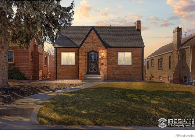 tudor-style house featuring entry steps, a lawn, brick siding, and roof with shingles