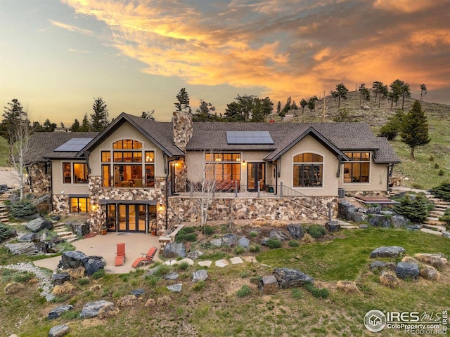 back of property at dusk with a patio area, roof mounted solar panels, stucco siding, a chimney, and stairway