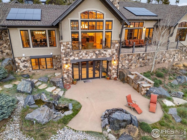 back of house featuring stucco siding, stone siding, roof mounted solar panels, a shingled roof, and a garage