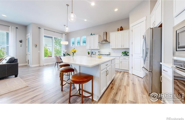 kitchen featuring stainless steel appliances, wall chimney exhaust hood, light wood-type flooring, a kitchen bar, and white cabinets