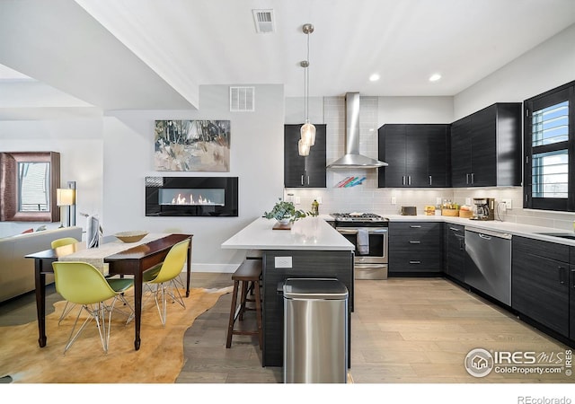 7861 W 51st Ave, Arvada, CO 80002 - kitchen featuring appliances with stainless steel finishes, wall chimney exhaust hood, dark cabinetry, light wood finished floors, and a kitchen breakfast bar