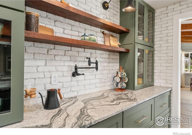 kitchen featuring green cabinets, open shelves, light stone counters, glass insert cabinets, and brick wall
