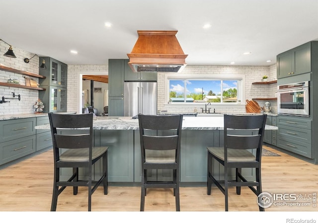 kitchen with stainless steel appliances, open shelves, brick wall, custom exhaust hood, and light stone countertops