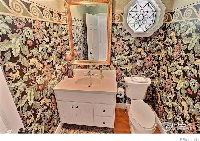 half bathroom featuring wallpapered walls, toilet, vanity, and wood finished floors