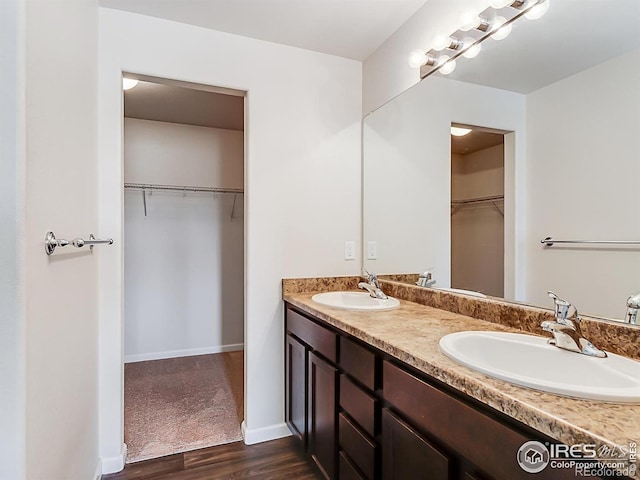 full bath with wood finished floors, double vanity, a walk in closet, and baseboards