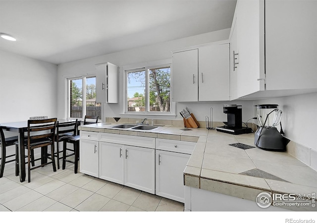kitchen with a sink, light tile patterned floors, tile counters, and white cabinets