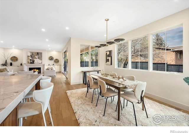 dining room with baseboards, a warm lit fireplace, recessed lighting, and light wood finished floors