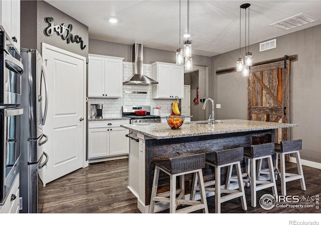 kitchen with wall chimney exhaust hood, a sink, stainless steel appliances, dark wood-type flooring, and an island with sink