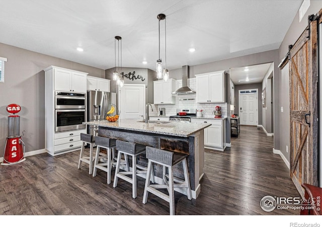 kitchen featuring wall chimney range hood, a barn door, stainless steel appliances, a sink, and a kitchen bar