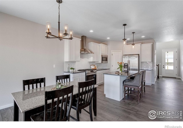 kitchen with appliances with stainless steel finishes, wall chimney exhaust hood, dark wood-style floors, a breakfast bar area, and an island with sink