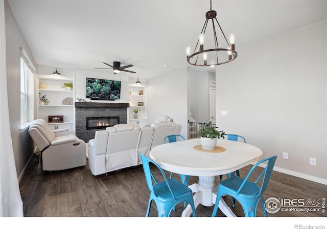 dining area with dark wood-style flooring, ceiling fan, baseboards, a glass covered fireplace, and a chandelier