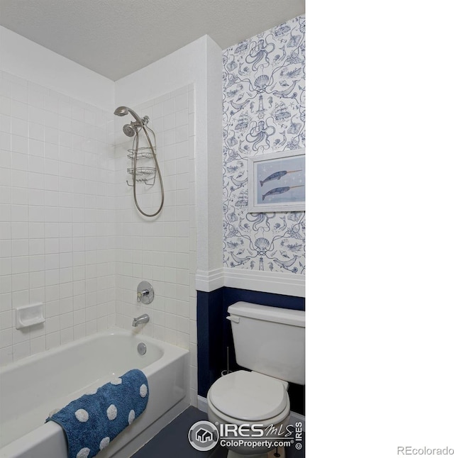 bathroom with wallpapered walls, toilet, wainscoting, and shower / washtub combination