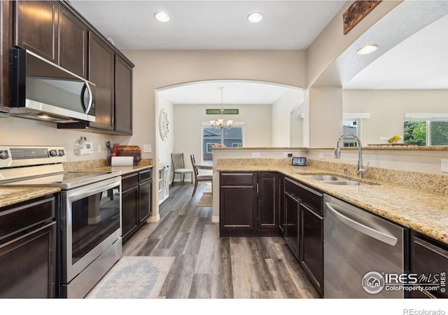 kitchen with stainless steel appliances, a sink, dark brown cabinetry, dark wood-style floors, and light stone countertops