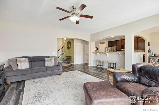 living area featuring arched walkways, dark wood-style flooring, a ceiling fan, stairway, and baseboards