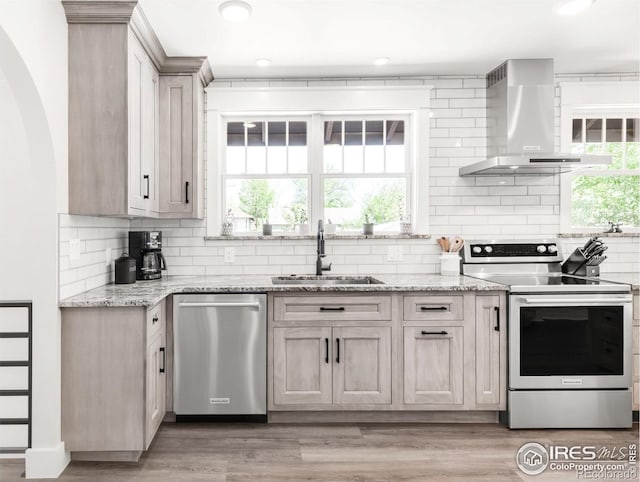 kitchen with stainless steel appliances, wall chimney exhaust hood, a sink, backsplash, and light stone counters