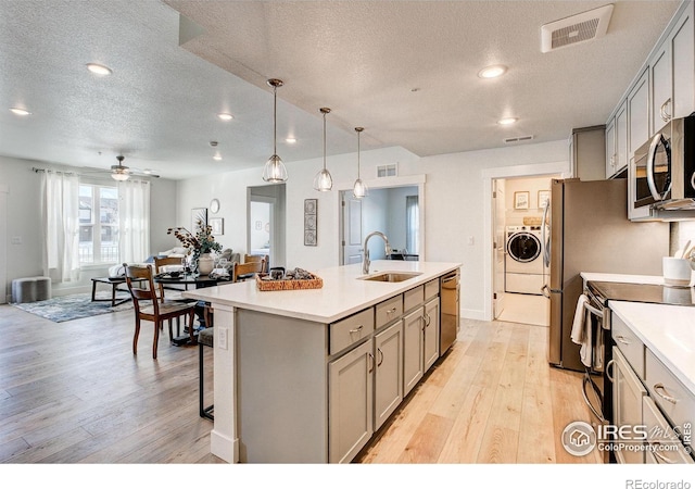 kitchen featuring stainless steel appliances, gray cabinetry, a textured ceiling, an island with sink, and light wood-style flooring