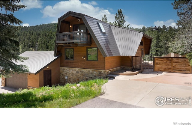 view of front of house featuring a balcony, a gate, a wooded view, stone siding, and metal roof