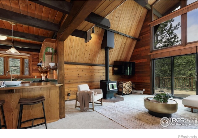 living area with wood ceiling, wooden walls, a wood stove, light colored carpet, and beamed ceiling