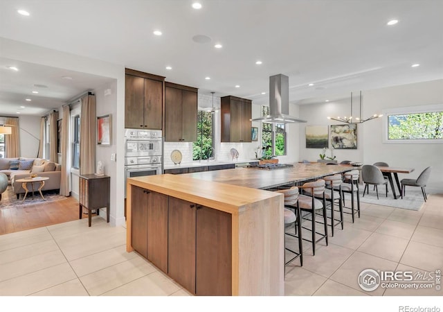 kitchen with light tile patterned floors, stainless steel double oven, healthy amount of natural light, island exhaust hood, and recessed lighting