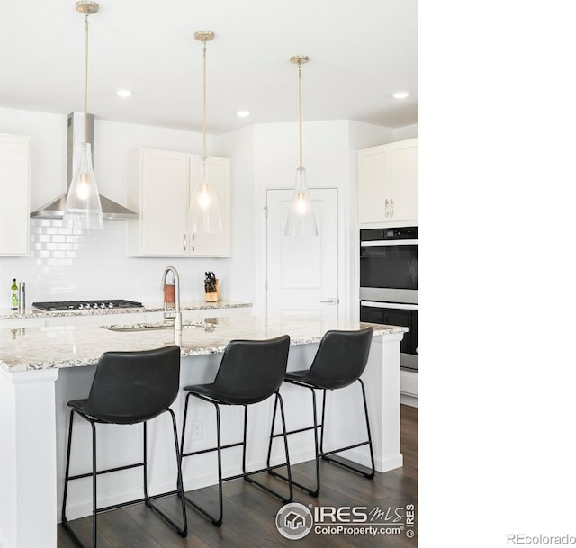 kitchen with wall chimney range hood, double wall oven, a sink, backsplash, and dark wood-type flooring