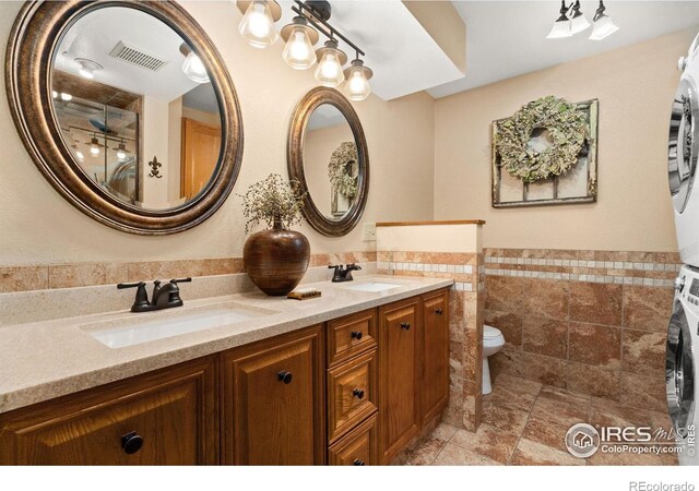 1738 S Estrella Ave, Loveland, CO 80537 - bathroom with stacked washer / drying machine, double vanity, tile walls, toilet, and a wainscoted wall