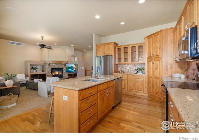 6787 Spanish Bay Dr, Windsor, CO 80550 - kitchen featuring stainless steel appliances, light wood finished floors, a kitchen island with sink, ceiling fan, and a glass covered fireplace