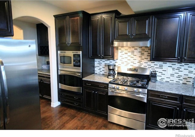 3106 Megan Way, Berthoud, CO 80513 - kitchen featuring appliances with stainless steel finishes, under cabinet range hood, arched walkways, dark wood-style floors, and decorative backsplash
