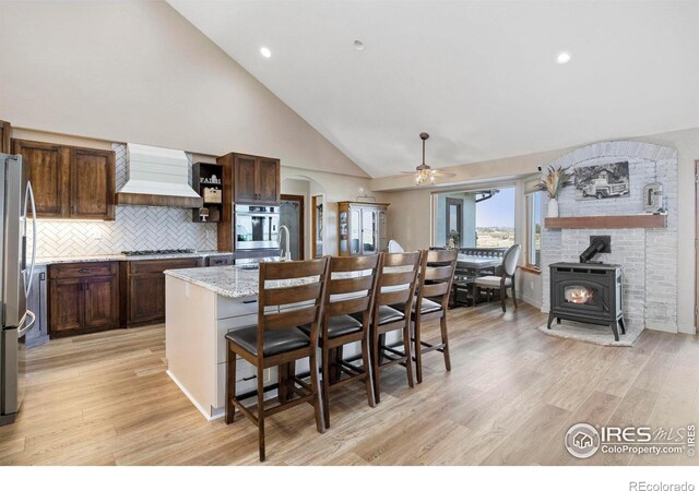 33820 County Road 47, Greeley, CO 80631 - kitchen featuring custom exhaust hood, a wood stove, appliances with stainless steel finishes, a center island with sink, and light wood-type flooring
