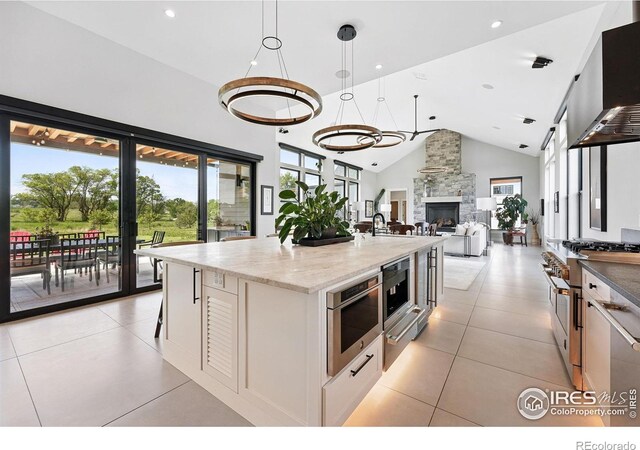 1563 Spring Creek Xing, Lafayette, CO 80026 - kitchen featuring wall chimney range hood, range with two ovens, a kitchen island with sink, light tile patterned flooring, and a stone fireplace