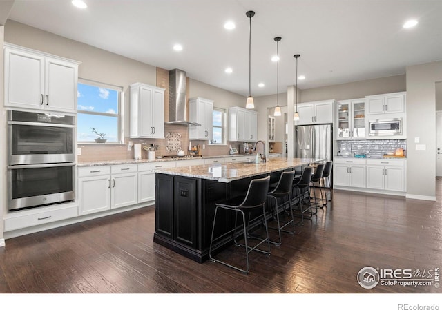 3075 Blue Mountain Dr, Broomfield, CO 80023 - kitchen featuring appliances with stainless steel finishes, wall chimney range hood, decorative backsplash, white cabinets, and a center island with sink