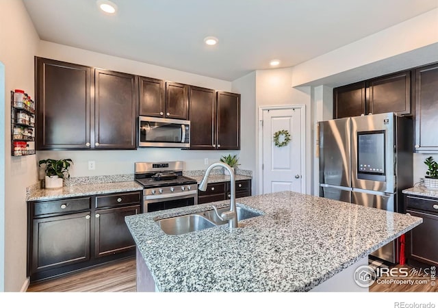 10 N Quantock St, Aurora, CO 80018 - kitchen with appliances with stainless steel finishes, light wood-style flooring, dark brown cabinetry, light stone countertops, and a kitchen island with sink