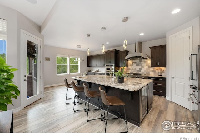 5966 Sunny Crest Dr, Timnath, CO 80547 - kitchen with wall chimney range hood, dark brown cabinetry, light wood-style flooring, backsplash, and a center island with sink