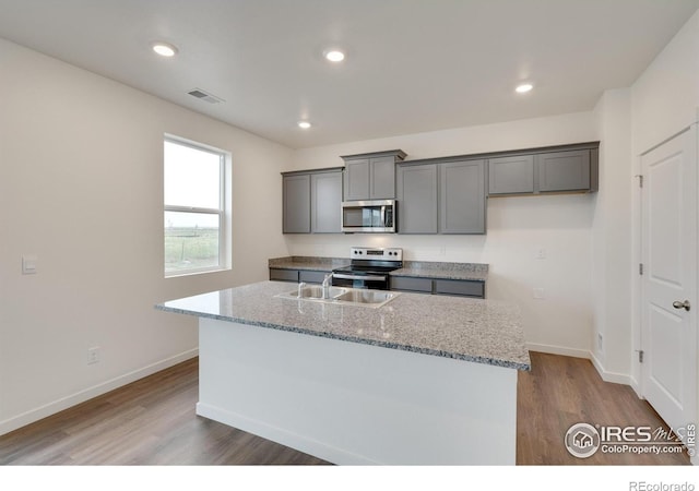 1639 Pinnacle Ave, Lochbuie, CO 80603 - kitchen featuring appliances with stainless steel finishes, gray cabinets, wood finished floors, light stone counters, and a kitchen island with sink