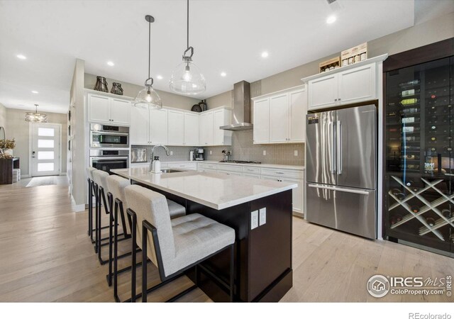 1768 Barefoot Dr, Windsor, CO 80550 - kitchen featuring appliances with stainless steel finishes, wall chimney exhaust hood, light wood-style flooring, tasteful backsplash, and an island with sink