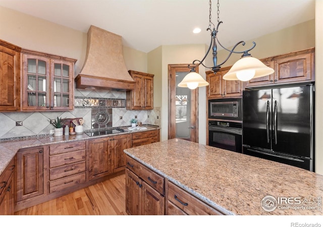 8276 Blackwood Dr, Windsor, CO 80550 - kitchen featuring black appliances, custom range hood, brown cabinets, light wood-style floors, and glass insert cabinets