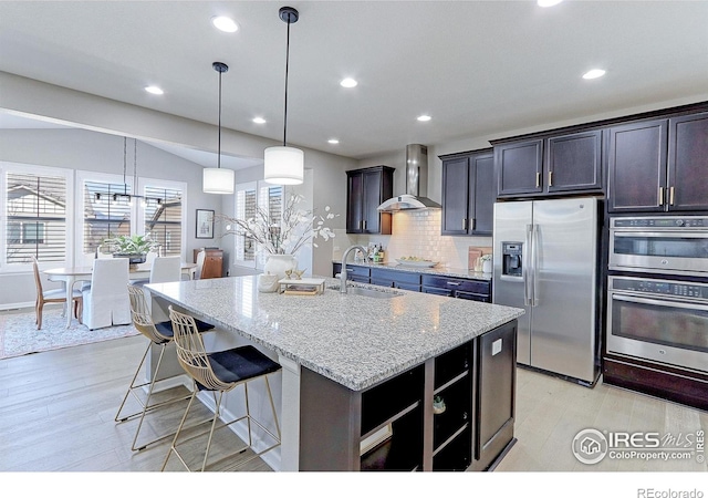 16021 Williams Pl, Broomfield, CO 80023 - kitchen featuring appliances with stainless steel finishes, wall chimney range hood, light stone countertops, light wood finished floors, and an island with sink