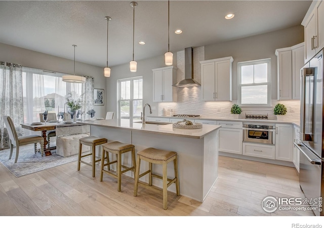 kitchen featuring appliances with stainless steel finishes, wall chimney exhaust hood, a center island with sink, and white cabinets