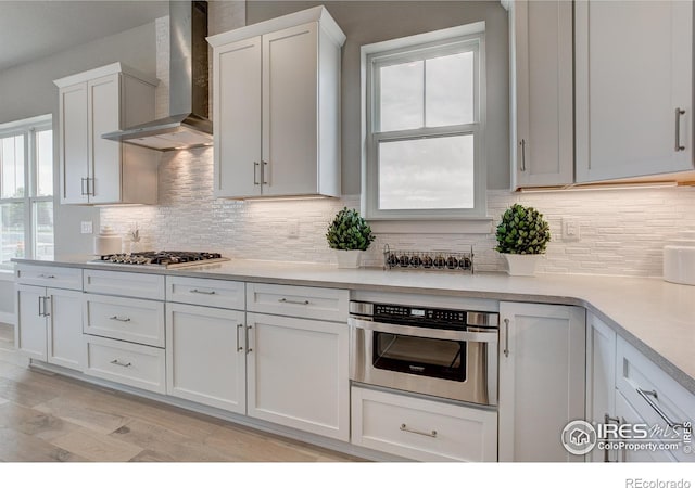 kitchen with light hardwood / wood-style flooring, appliances with stainless steel finishes, white cabinets, wall chimney range hood, and backsplash