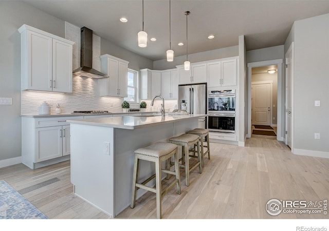 kitchen featuring sink, a kitchen island with sink, white cabinetry, stainless steel appliances, and wall chimney exhaust hood