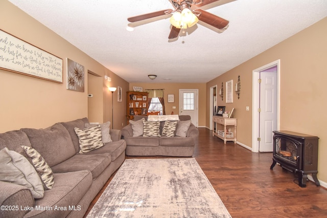 living room featuring a textured ceiling, dark wood-style floors, baseboards, ceiling fan, and a wood stove