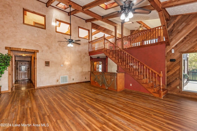 unfurnished living room featuring wood walls, ceiling fan, a towering ceiling, beamed ceiling, and wood-type flooring